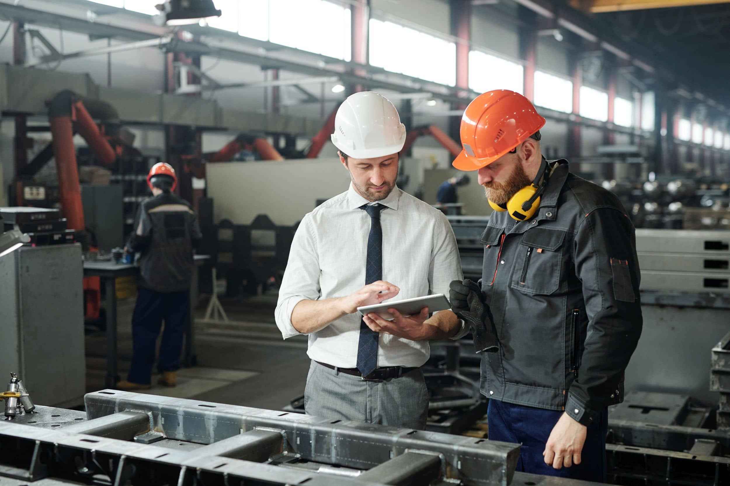 Fokusbranche Maschinenbau und Metallverarbeitung, Baustein Sachversicherung: 2 Mitarbeitende mit Helm in Fabrikhalle konfigurieren eine Maschine.