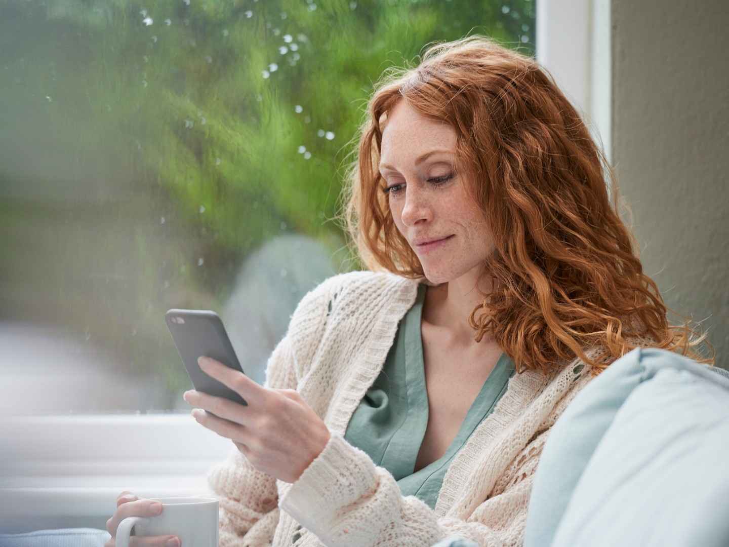 Eine junge Frau trinkt einen Kaffee und schaut dabei auf ihr Handy. 