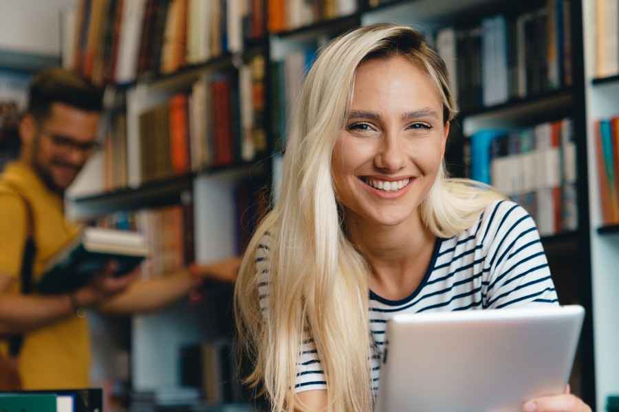 Eine junge Frau sitzt in einer Bibliothek und lächelt in die Kamera.